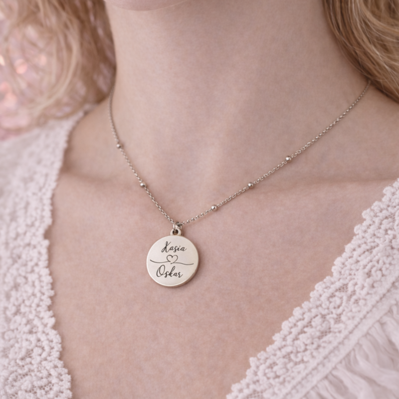 Srebrny personalizowany naszyjnik z okrągłą zawieszką 2 cm zaprezentowany na szyi modelki. Delikatny łańcuszek i subtelny grawer imion podkreślają lekki, elegancki charakter biżuterii.